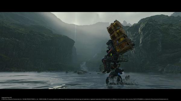 Screenshot from DEATH STRANDING DIRECTOR'S CUT showing Sam Bridges carrying cargo through a river in a lush, mountainous landscape.