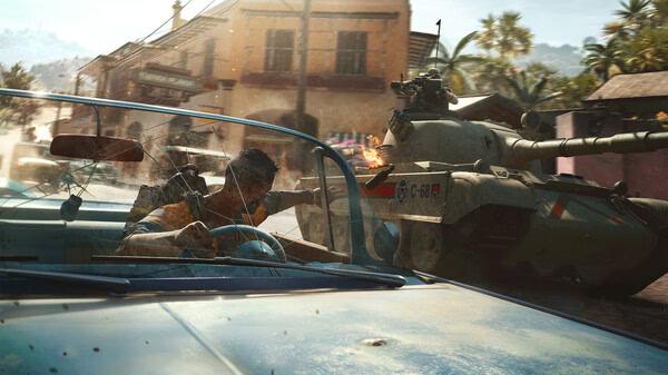 A screenshot from Far Cry® 6 showing a car chase with a man driving while a tank fires in a vibrant, tropical town setting.