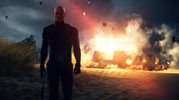 HITMAN World of Assassination screenshot: A figure with a weapon stands before a fiery explosion on a desolate beach at dusk.