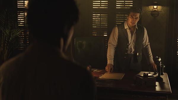 Screenshot from Mafia: The Old Country showing a tense meeting in a dimly lit office with two men discussing at a wooden desk.
