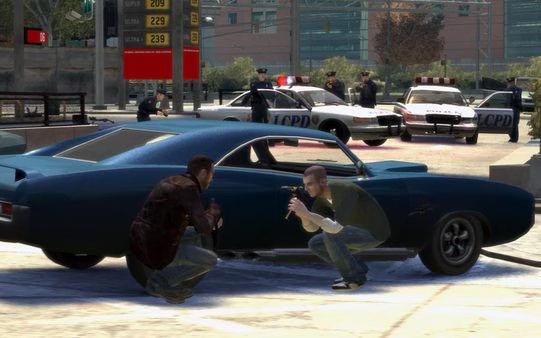 Screenshot from Grand Theft Auto IV showing two characters crouched by a blue car with police cars and officers nearby.