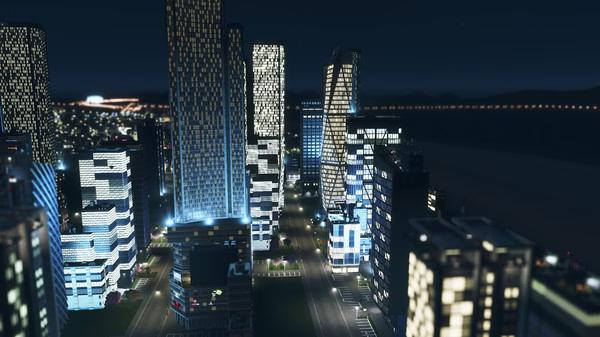 A nighttime screenshot from Cities: Skylines showing a brightly lit cityscape with modern skyscrapers and a river in the background.