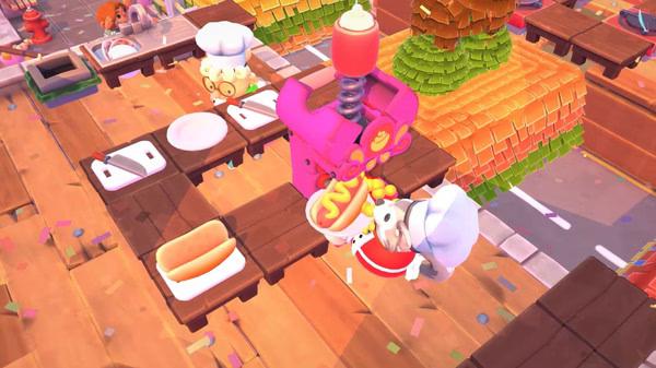 Overcooked! 2 screenshot: Chefs prepare hot dogs using a condiment dispenser in a colorful kitchen with scattered confetti.