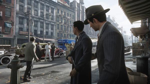 Screenshot from Mafia: Definitive Edition showing two men in suits observing a street scene with vintage cars and buildings.