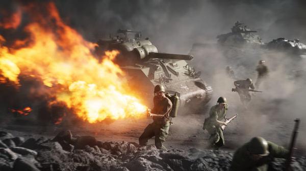 Battlefield V screenshot: Soldiers advance under fiery explosions, with tanks visible in the background amidst smoke and debris.