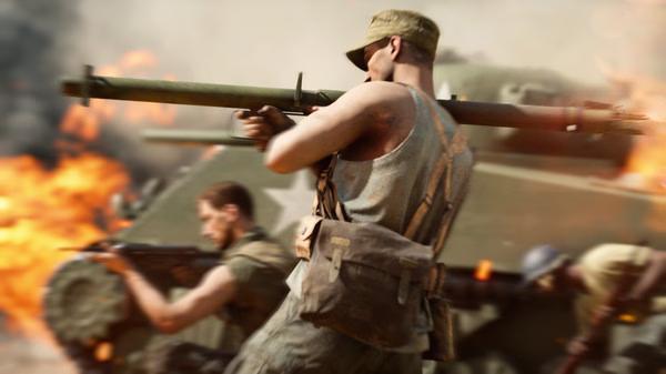 Battlefield V screenshot: Soldiers in combat gear advance with a bazooka, tank and fire in the background.