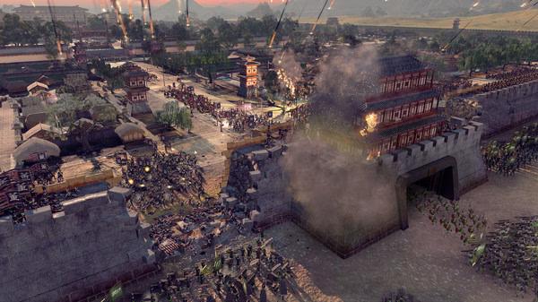 Screenshot from Total War: Three Kingdoms showing a dramatic siege with troops attacking a fortified city under heavy fire.