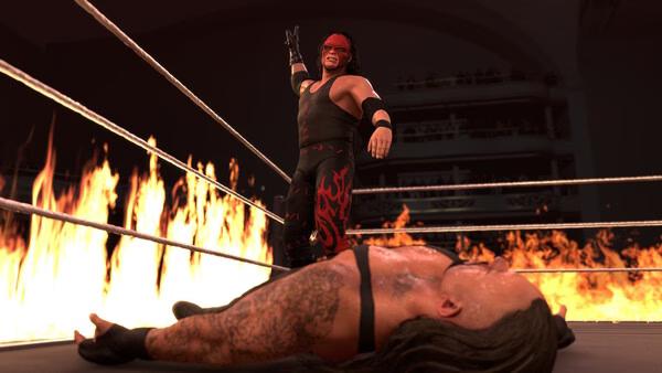WWE 2K26 screenshot: A wrestler in a black and red outfit stands triumphantly over a fallen opponent in a ring surrounded by flames.