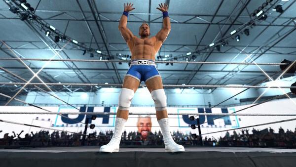 WWE 2K26 screenshot: Wrestler in blue trunks and white boots stands triumphantly in the ring, arms raised, with a cheering crowd behind.