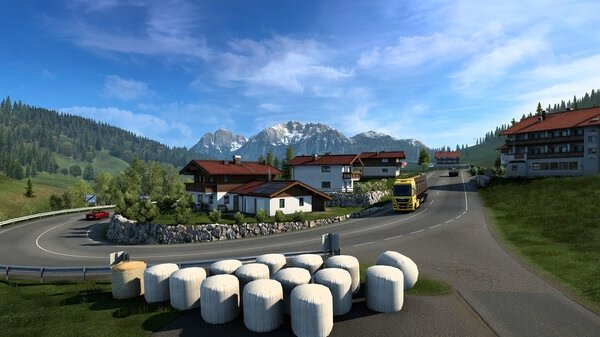 A screenshot from Euro Truck Simulator 2 showing a truck driving past houses in a scenic mountain area with hay bales in the foreground.