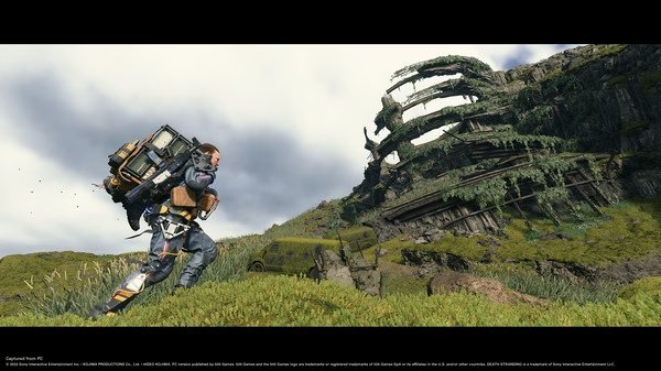 Screenshot from DEATH STRANDING DIRECTOR'S CUT shows a character carrying a large backpack, walking towards a grassy, curved structure.
