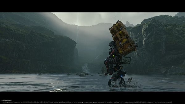 Screenshot from DEATH STRANDING DIRECTOR'S CUT showing Sam Bridges carrying cargo through a river in a lush, mountainous landscape.