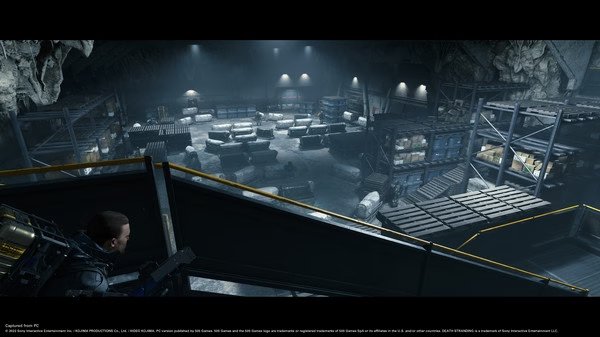 Screenshot from DEATH STRANDING DIRECTOR'S CUT showing a character overlooking a large, futuristic warehouse filled with cargo and storage units.
