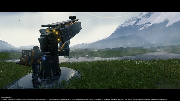Screenshot from DEATH STRANDING DIRECTOR'S CUT showing a character standing beside a large cargo-launching device in a grassy landscape.