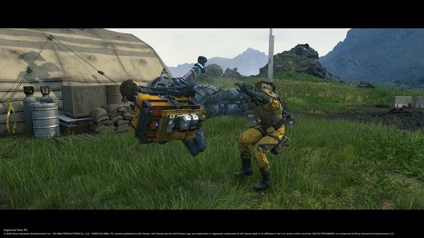 Screenshot from DEATH STRANDING DIRECTOR'S CUT showing a character in yellow gear lifting another character near a tent in a grassy landscape.