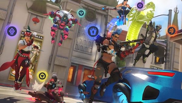 Overwatch® screenshot showing a dynamic battle scene with multiple heroes in action, each marked by colorful icons, against a city backdrop.