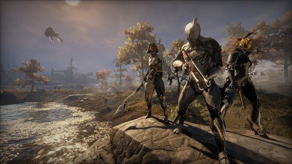Three Warframe characters stand on a rock by a river, armed and ready, under a bright sky. Warframe game screenshot.