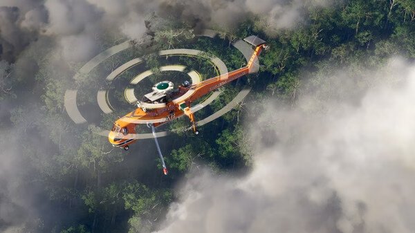 Screenshot from Microsoft Flight Simulator 2024 showing an orange helicopter flying over a dense forest below misty clouds.