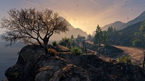 A scenic view in Grand Theft Auto V shows a sunset over mountains and a winding road. Screenshot includes a tree on a rocky cliff.