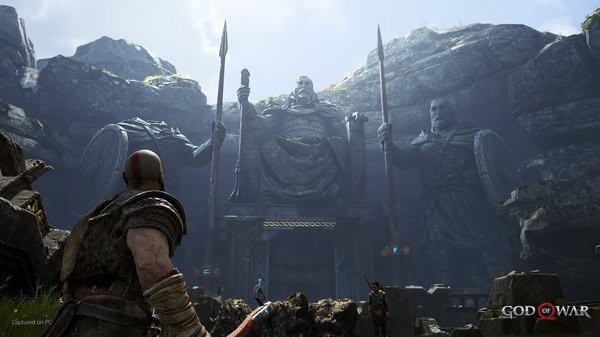 Kratos stands before a towering statue of Norse gods in a rocky landscape in this 'God of War' screenshot.