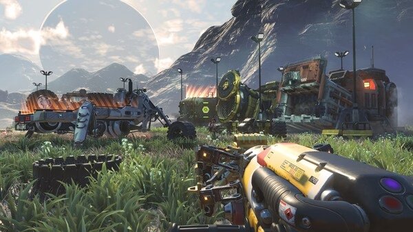 Screenshot from No Man's Sky showing a futuristic base with vehicles and machinery against a mountainous landscape.